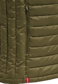 Olivgrüner gesteppter Jackenabschnitt mit horizontalen Nähten, Reißverschlusstasche und kleinem roten Stoffmarkenetikett nahe dem Saum.