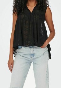 Musta hihaton paita, jossa on laskostettu etuosa, puoliksi läpinäkyvä kangas ja V-pääntie, yhdistettynä vaaleansinihin korkeavyötäröisiin farkkuihin.