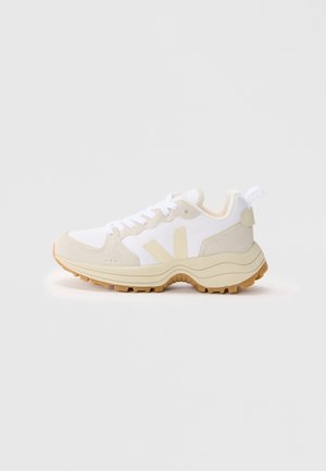 White and beige sneaker with mesh upper, suede accents, and textured rubber sole. Features a rear tab and stylized side logo.