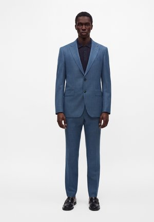 Homme portant un costume bleu ajusté avec une veste à deux boutons, un polo noir et des chaussures habillées noires, debout devant un fond uni.