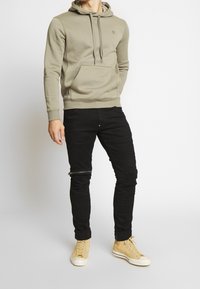 Olivgrön hoodie med ficka och dragsnören, kombinerad med svarta skinny jeans med dragkedjor vid knäna, bärs med beige sneakers.