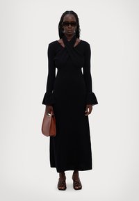 Robe noire à manches longues avec un devant croisé, des manches évasées et une silhouette ajustée. Assortie à un sac à main marron et des sandales noires à lanières.