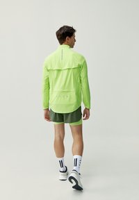 Giacca leggera a maniche lunghe di colore verde neon con dettagli riflettenti, abbinata a pantaloni corti di colore verde oliva. Tessuto testurizzato, dettagli delle cuciture minimali.
