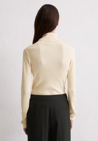 Pull en maille côtelée jaune clair avec col roulé et manches longues, associé à des pantalons noirs taille haute, montré de dos.