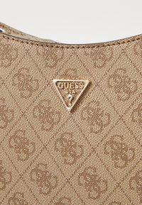 Beige textúrájú kézitáska, amelyen egy háromszög alakú, aranyszínű "GUESS USA" logó és egy ismétlődő minta található a felületen.
