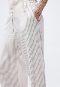 Ανοιχτόχρωμο παντελόνι με ψιλές ρίγες, με σχέδιο που στενεύει προς τα κάτω και ορατές πλαϊνές τσέπες. Το ύφασμα φαίνεται λείο με διακριτική γυαλάδα.