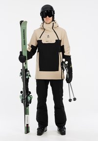 Ski-kleding met een beige en zwarte jas met een hoge kraag, zwarte broek, zwarte handschoenen en groene ski's met bindingen.