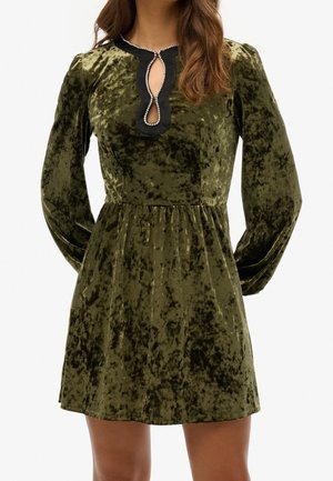 Kobieta w oliwkowej sukience z marszczonego aksamitu z długimi bufiastymi rękawami i czarnym dekoltem w kształcie łezki wykończonym perełkami.