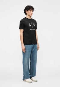 Svart t-skjorte i bomull med hvitt logoavtrykk; kombinert med lyseblå bukser med vide ben og hvite joggesko. Enkel, avslappet design.