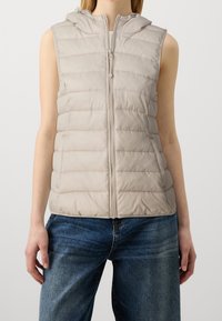 Veste matelassée beige clair avec capuche, dotée d'une fermeture éclair frontale et d'un motif matelassé vertical, associée à un pantalon en denim bleu foncé.