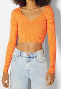 Pomarańczowy, prążkowany, długi rękaw crop top z dekoltem w kształcie litery V, w zestawie z wysokim pasem jasnoniebieskimi dżinsami. Gładka tekstura z dopasowanym krojem.