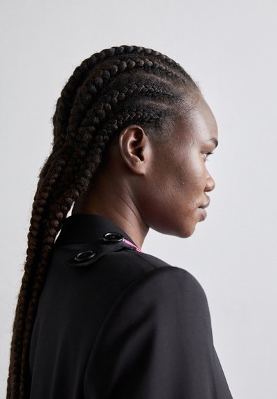 Veste noire ajustée avec un col haut, arborant deux boutons ronds noirs et des accents roses, portée par un modèle avec de longues tresses.