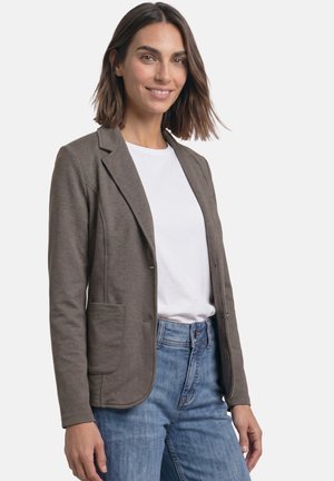 Kvinde smiler, iført en brun blazer over en hvid skjorte og blå jeans, står mod en ensfarvet lys baggrund.