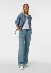 Jasnoniebieska kurtka dżinsowa z zapięciem na guziki i naszywaną kieszenią, w zestawie z dopasowanymi, luźnymi dżinsami oraz białymi trampkami. Prosty design.