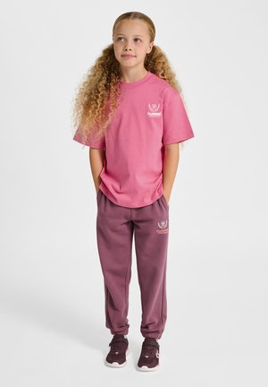 Młoda dziewczyna z długimi kręconymi włosami, ubrana w różowy t-shirt hummel, fioletowe spodnie dresowe i ciemnofioletowe sneakersy, stojąca z rękami w kieszeniach.