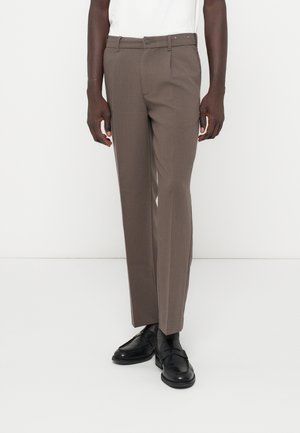 Homme portant un pantalon droit taupe, des mocassins en cuir noir et une chemise blanche, debout devant un fond blanc uni.