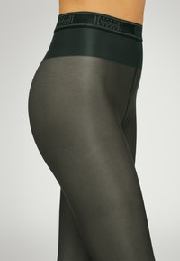 Collant trasparente verde scuro con un girovita opaco e liscio che presenta il logo Wolford. Il tessuto ha una finitura lucida e una texture morbida.