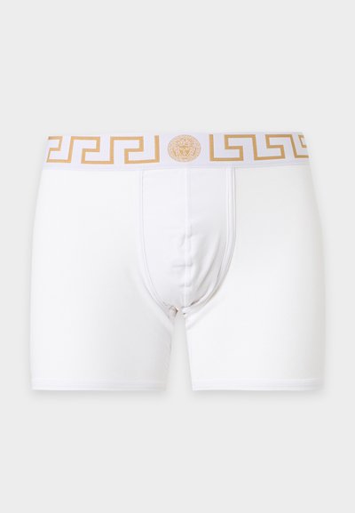 Boxers blancs pour hommes avec un motif clé grec doré et un logo Méduse sur la ceinture, sur fond gris.