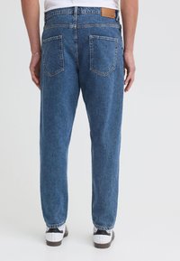 Homme portant un jean bleu ample, des baskets blanches et une chemise blanche, vu de dos sur un fond uni.