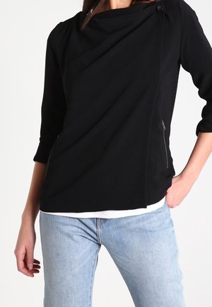 Femme portant un haut noir asymétrique zippé avec manches trois-quarts et un jean bleu clair, main dans la poche sur fond blanc.
