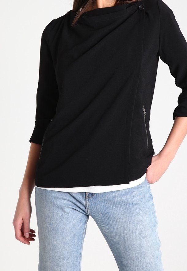 Femme portant un haut noir asymétrique zippé avec manches trois-quarts et un jean bleu clair, main dans la poche sur fond blanc.