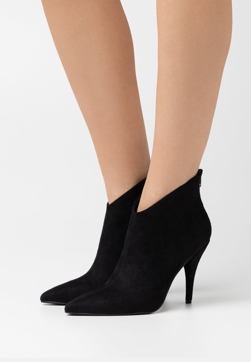 Bottines en daim noir à bout pointu et talons hauts portées sur jambes nues, avec des talons aiguilles fins et une fermeture éclair à l'arrière.