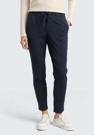 Persona con pantalones azules marinos ajustados con cintura de cordón y zapatillas blancas, de pie con una mano en el bolsillo.