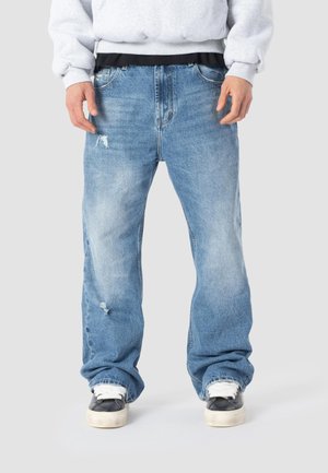 Hombre con jeans holgados desgastados de color azul claro, zapatillas blancas y negras y sudadera gris clara, de pie frente a un fondo liso.