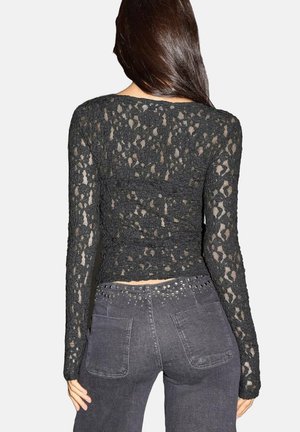 Schwarzes Spitzenoberteil mit langen Ärmeln, das ein tailliertes Design mit floralem Muster und einer cropped Länge aufweist, kombiniert mit dunkelgrauen Jeans.