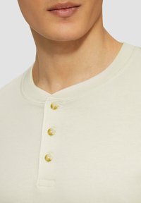 Cremefarbendes Henley-Shirt mit Rundhalsausschnitt, gerippter Textur und drei falschen Perlmuttknöpfen an der Knopfleiste.