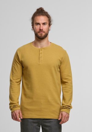 Bearded man mit zurückgebundenem Haar, der ein senfgelbliches Langarm-Henley-Shirt und graue Hosen trägt und vor einem einfachen hellgrauen Hintergrund steht.