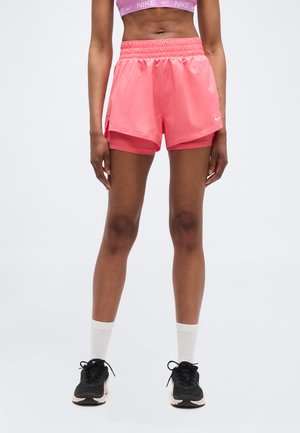 Femme portant un soutien-gorge de sport Nike rose et un short à couches assorti, avec des chaussures de course noires et des chaussettes blanches, debout devant un fond uni.