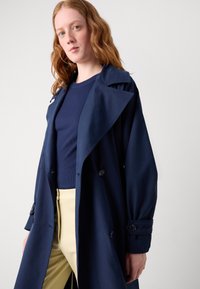 Manteau trench bleu marine avec un design à double boutonnage, larges revers et poignets boutonnés, porté par-dessus un haut bleu marine et un pantalon jaune.