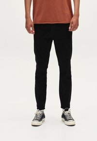Crne slim-fit hlače od pamuka, uparene s tamnoplave platnene tenisice s bijelim vezicama i potplatima. Košulja je boje hrđe.