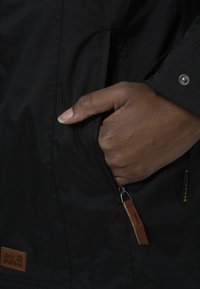 Black waterproof jacket with a side zipper; leather pull tab; fabric has a smooth texture; logo patch near the bottom left.