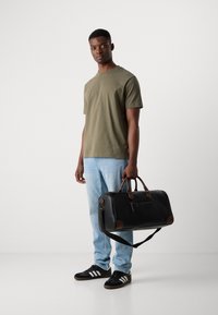 Black leather duffel bag with brown accents, zipper, and adjustable strap. Model wears olive t-shirt and light blue jeans, holding the bag.