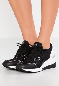 Svart och vit atletisk sko med en texturerad ovandel, snören och en glansig finish på tån. Sklisäker gummisula med en lätt klack.