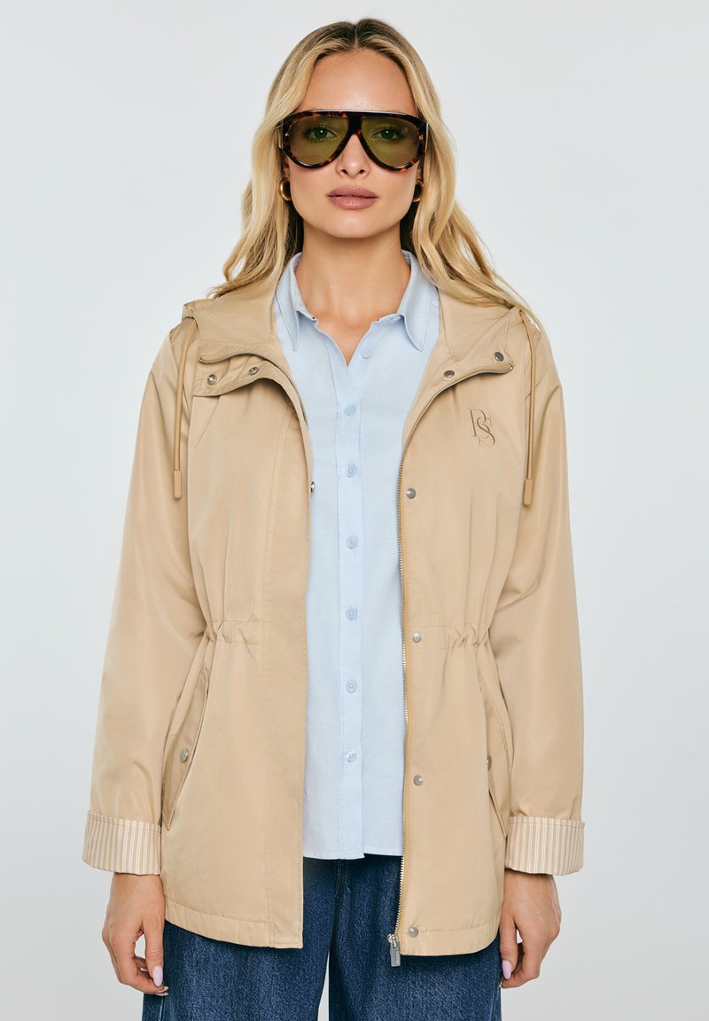 Naine, kes kannab beeži tõmblukuga jopet helesinise nööbitava särgi kohal, suured toonitud päikeseprillid, lahtised blondid juuksed ja üleskääritud varrukad.