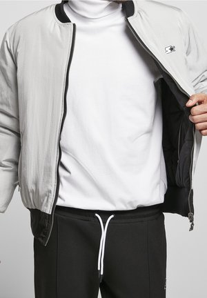 Homme portant une veste bomber gris clair ouverte sur un col roulé blanc et un pantalon noir à cordon, sur un fond uni.