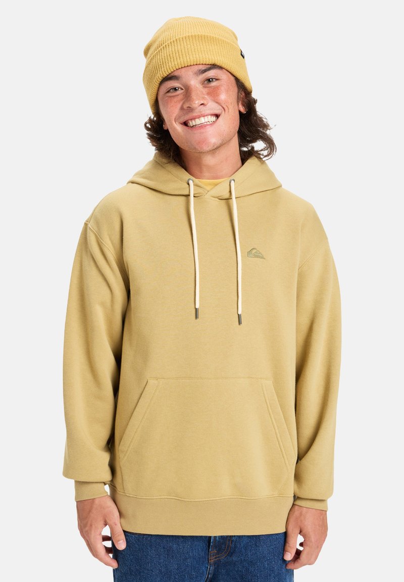 Sudadera caqui con un bolsillo frontal, capucha con cordón y un pequeño logo en el pecho. Combinada con un gorro amarillo de canalé.