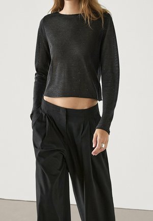 Femme portant un crop top gris foncé ajusté à manches longues et un pantalon noir ample taille haute, avec la main gauche dans la poche.