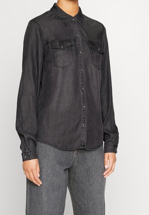Person iført en sort langærmet denim skjorte med brystlommer og knapper samt grå jeans, stående mod en ensfarvet baggrund.