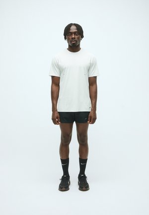 T-shirt gris clair à manches courtes avec des accents noirs, associé à un short noir, des chaussettes noires et des chaussures de sport noires, se tenant devant un fond uni.