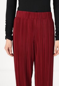 Red pleated pants with a high elastic waistband. The fabric features vertical pinstripes, providing texture and structure.