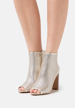 Kovově stříbrné kotníkové boty s perforovaným designem, otevřenou špičkou a zapínáním na zip. Podpatek typu block a hladká textura.