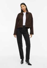 Brūna, teksturēta īsa jaka ar apkakli un divām priekšējām kabatām, valkāta pāri baltai kreklam kopā ar augstajās vidukļa melnajām džinsu biksēm un melniem papēžiem.