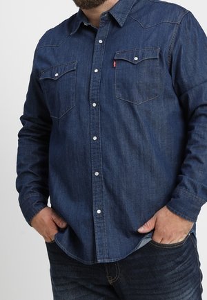 Homme portant une chemise en denim bleu foncé avec boutons et poches à rabat, mains dans les poches, visible du cou jusqu’aux hanches.