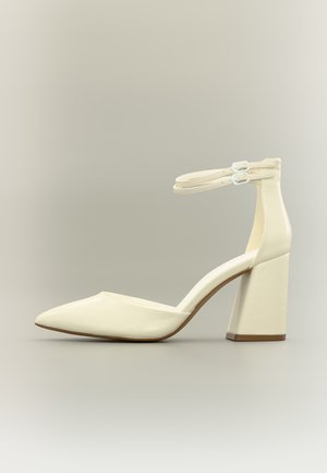 Chaussure à talon haut crème à bout pointu avec doubles brides à la cheville et talon bloc sur fond neutre.