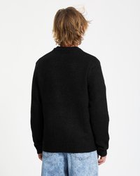 Person with shoulder-length blond hair wears black sweater and light blue acid-wash jeans, standing facing a white background.