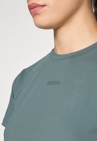 Primeiro plano de uma pessoa a usar uma t-shirt verde suave com "BORG" impresso no peito, mostrando o pescoço, a linha do maxilar e a parte superior do torso.